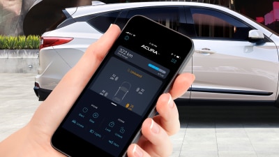 Closeup of hand holding a phone with AcuraLink app open on screen. In the background, a white Acura RDX parked in front of modern architectural building.