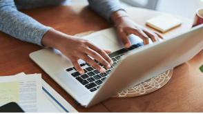 Closeup bird’s eye view of a woman’s hands, typing on her laptop at a wooden table.