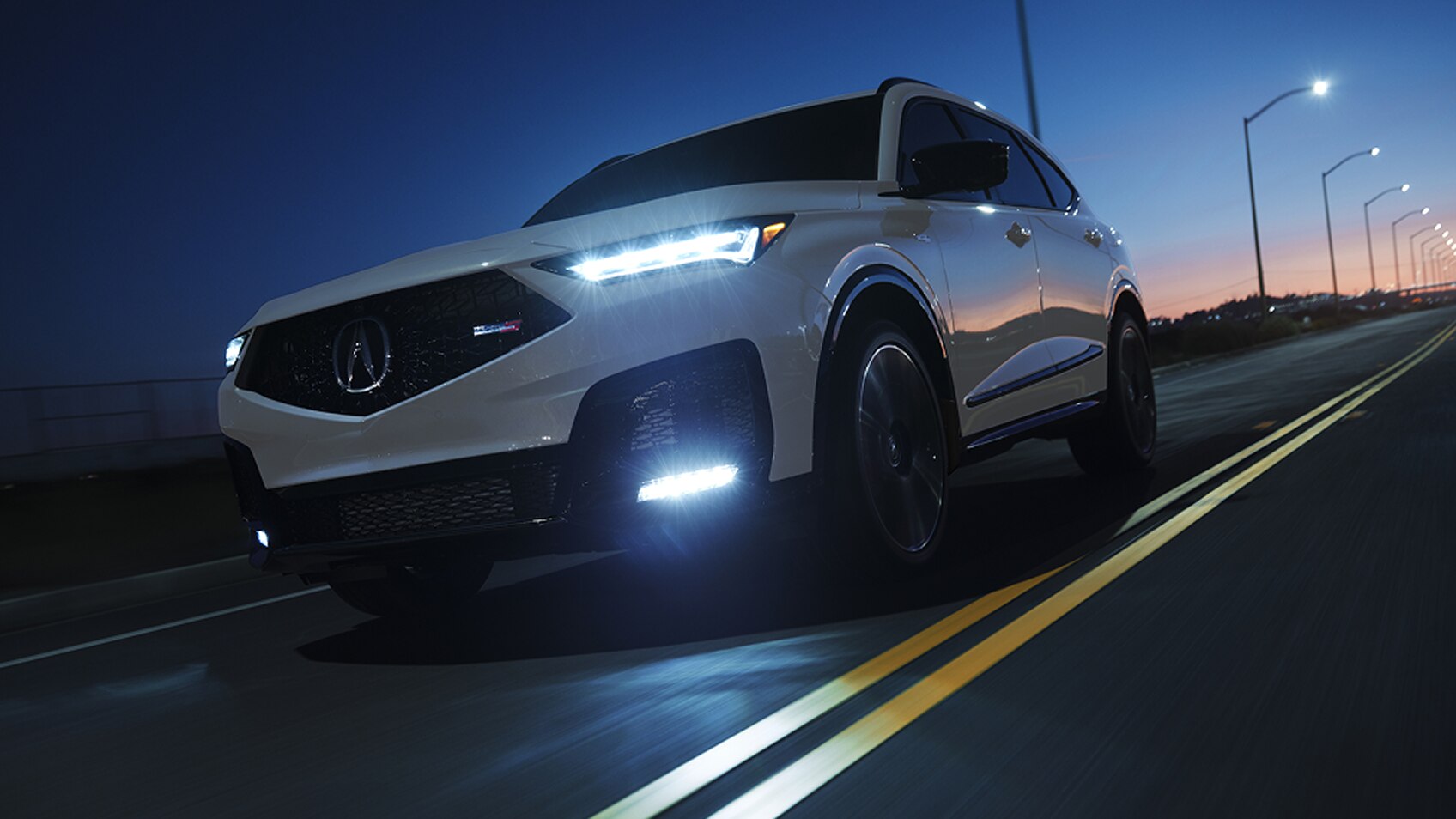 Un MDX Type S 2025 blanc roule de nuit, phares allumés, sur une autoroute éclairée.