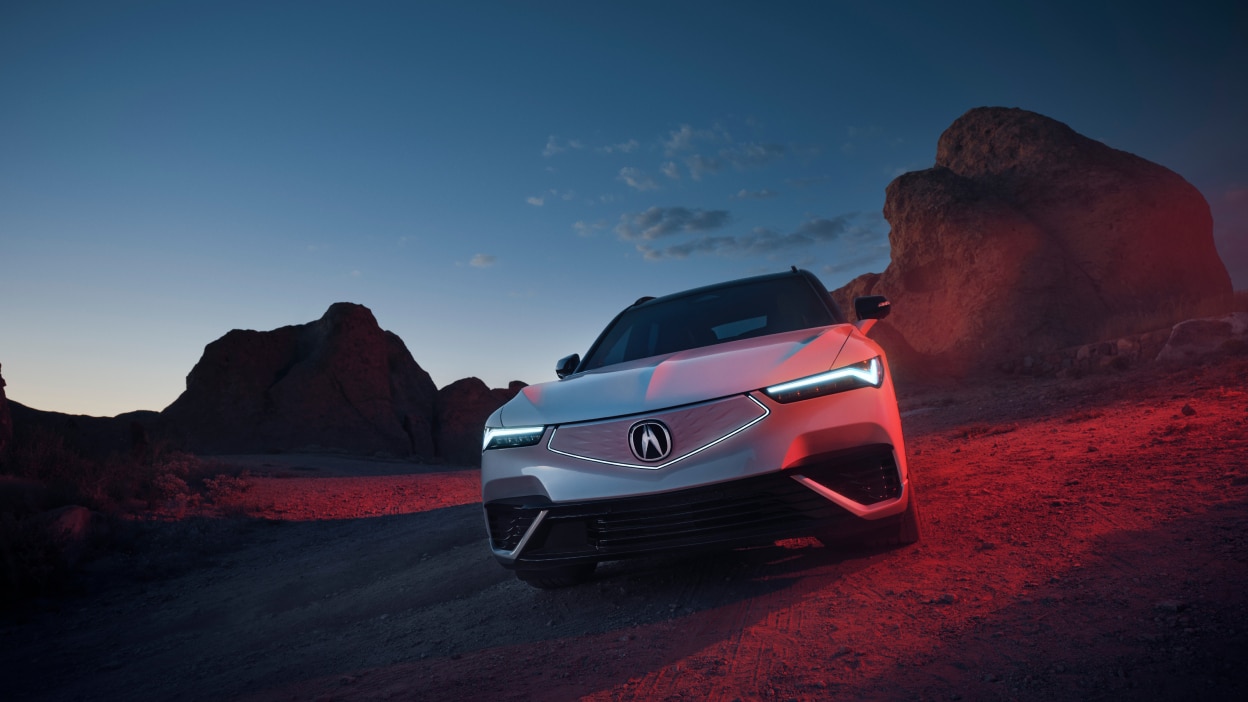 Medium wide front view of white ZDX parked in the desert at dusk. Its headlights are on and the diamond pentagon front fascia is illuminated.