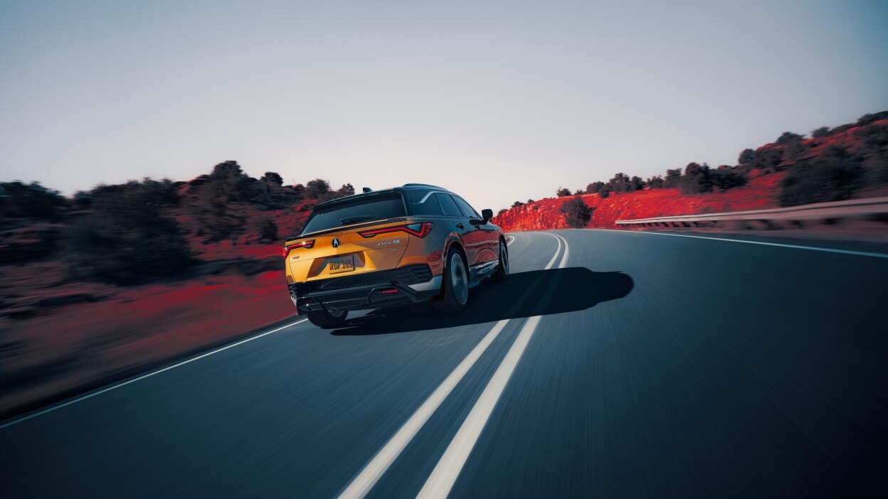 3/4 rear view of gold ZDX driving on mountain highway. Part of the image is saturated with colour, so the dirt on the sides of the highway is red.