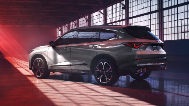 Plan de trois quarts latéral arrière d’un MDX gris dans un entrepôt vide, éclairé partiellement par la lumière naturelle et partiellement par la lumière rouge.