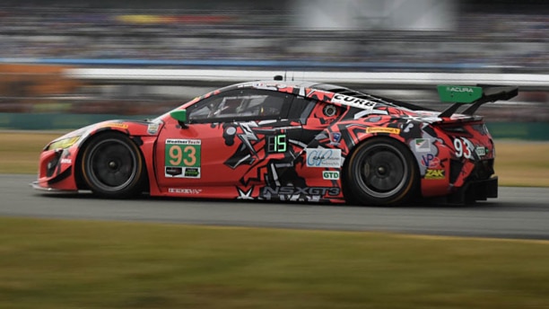 Side view of a red Acura NSX GT3 wrapped in sponsorship logos, driving at high speed on a racetrack, the stad ium blurred in the background. Vue latérale d’une Acura NSX GT3 rouge recouverte de logos de commanditaires roulant à haute vitesse sur un circuit de course avec des estrades en arrière-plan.