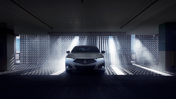 Front view of a white Acura TLX in a darkened garage with natural light shining through a gated wall behind. // Vue avant d’une Acura TLX blanche dans un garage sombre devant un mur qui laisse passer la lumière naturelle.