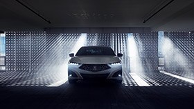 Front view of a white Acura TLX in a darkened garage with natural light shining through a gated wall behind. // Vue avant d’une Acura TLX blanche dans un garage sombre devant un mur qui laisse passer la lumière naturelle.