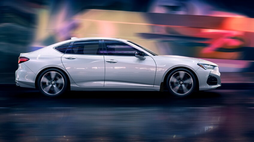 Passenger side view of a white Acura TLX driving at night - red, purple and yellow lights are blurred in the background by the motion of the car. // Vue du côté passager d’une Acura TLX blanche circulant la nuit avec en arrière-plan des lumières floues rouges, violettes et jaunes.