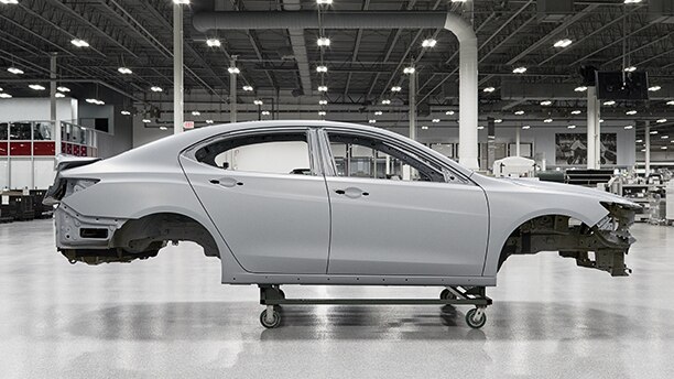Passenger side view of a white vehicle frame hoisted on a dolly inside the Acura Performance Manufacturing Centre	Vue du côté passager d’un cadre de véhicule blanc sur un chariot à l’intérieur du Performance Manufacturing Centre d’Acura.
